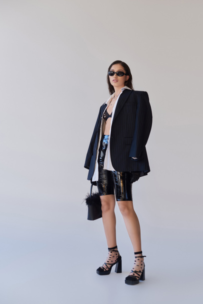 divatos megjelenés, divat nyilatkozat, barna ázsiai nő napszemüvegben pózol tollas táska szürke háttér, modell latex rövidnadrág, melltartó, fekete kabát és kesztyű, fiatalok, teljes hossz  - Fotó, kép