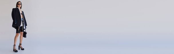 Trendlook, Mode nach vorne, brünette asiatische Frau mit Sonnenbrille posiert mit gefiederter Handtasche auf grauem Hintergrund, Model in Latex-Shorts, BH, schwarzer Jacke und Handschuhen, Jugend, volle Länge, Banner - Foto, Bild