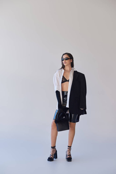 cool style, studio photography, young asian woman in stylish look and sunglasses posing with feathered handbag on grey background, blazer and latex shorts, youthful fashion, full length  - Foto, imagen