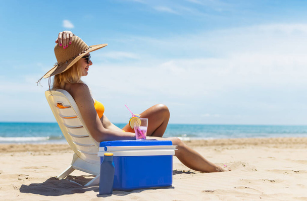 Feliz joven europea bebe un cóctel y descansa en una tumbona en la playa, espacio para una inscripción portátil refrigerador azul junto al mar. Foto de alta calidad - Foto, Imagen