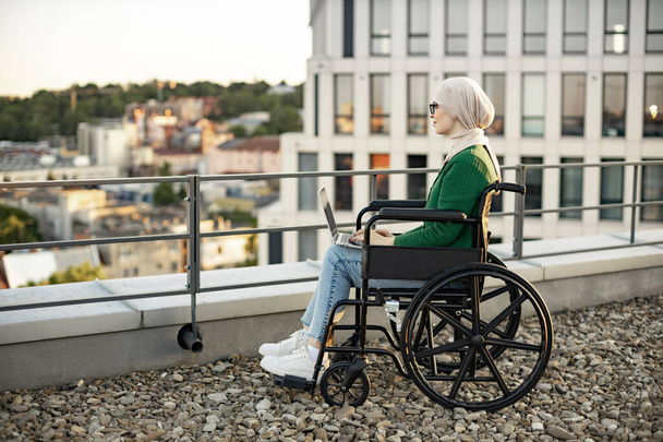 Side view of muslim lady in headscarf using digital device for remote work on panoramic terrace. Well -organized businesswoman in wheelchair searching information over internet application outdoors. - Fotoğraf, Görsel