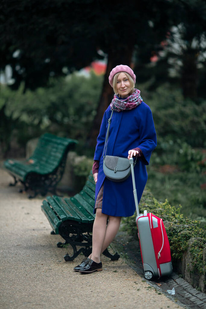 A woman with a red suitcase is standing in the park, next to a bench. - Fotoğraf, Görsel