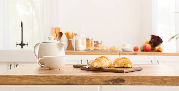 Leikkuulauta croissanteilla, veitsellä, teekannulla ja kupilla puisella pöydällä modernissa keittiössä - Valokuva, kuva