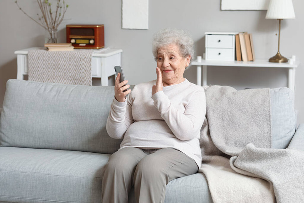 Femme âgée avec téléphone portable vidéo bavardage sur canapé à la maison - Photo, image