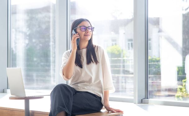 Χαρούμενη νεαρή γυναίκα με γυαλιά μιλώντας σε ένα smartphone στο γραφείο στο παρασκήνιο του παραθύρου, η έννοια της εργασίας. - Φωτογραφία, εικόνα