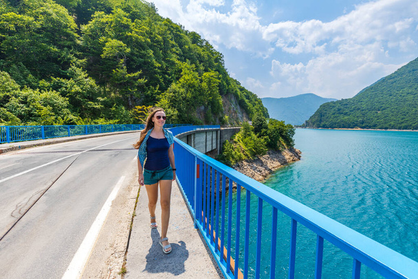 Young woman walking on the bridge across Piva lake in Montenegro. Female tourist spending time on vacations near the lake - Foto, afbeelding