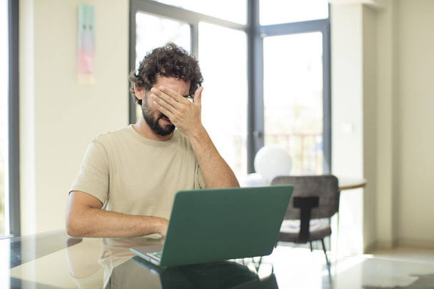 young adult bearded man with a laptop covering eyes with one hand feeling scared or anxious, wondering or blindly waiting for a surprise - Foto, Imagem
