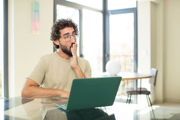 young adult bearded man with a laptop yawning lazily early in the morning, waking and looking sleepy, tired and bored - 写真・画像