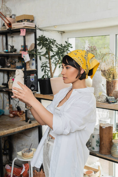 Νεαρή μελαχρινή Ασιάτισσα καλλιτέχνης με μαντίλα και ενδύματα εργασίας κρατώντας πήλινο προϊόν, ενώ στέκεται σε θολή τάξη κεραμικής στο παρασκήνιο, στούντιο κεραμικής με τεχνίτη στην εργασία - Φωτογραφία, εικόνα