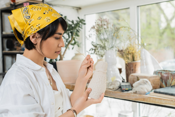 Young asian brunette artist in headscarf and workwear holding clay sculpture and wooden stick while working in blurred pottery class at background, pottery studio with artisan at work - Photo, Image
