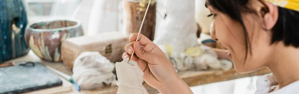 Oldalnézet fiatal mosolygós elmosódott ázsiai művész kezében fa bottal és agyag szobor, miközben dolgozik elmosódott fazekas osztály a háttérben, fazekas stúdió kézműves a munkahelyen, banner  - Fotó, kép