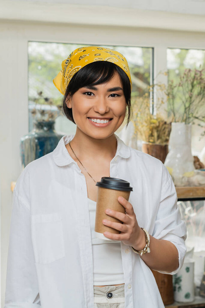 Portrait of smiling young asian female artisan in headscarf and workwear looking at camera and holding coffee to go while standing in blurred pottery class, pottery workshop with skilled artisan - Foto, immagini