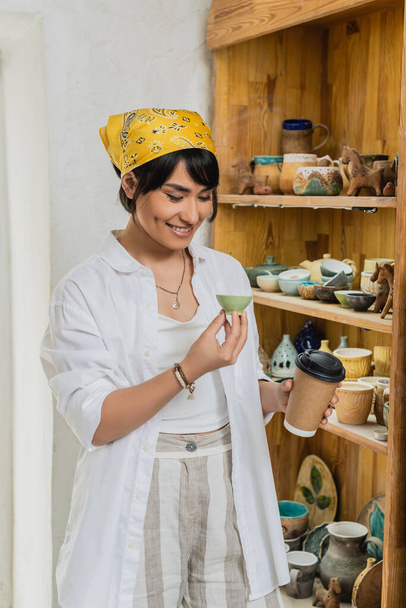 Χαρούμενος νεαρός Ασιάτης καλλιτέχνης με μαντίλα και ενδύματα εργασίας κρατώντας καφέ για να πάει και πήλινο προϊόν και στέκεται κοντά σε ράφια με γλυπτά στο παρασκήνιο, εργαστήριο κεραμικής με εξειδικευμένο τεχνίτη - Φωτογραφία, εικόνα