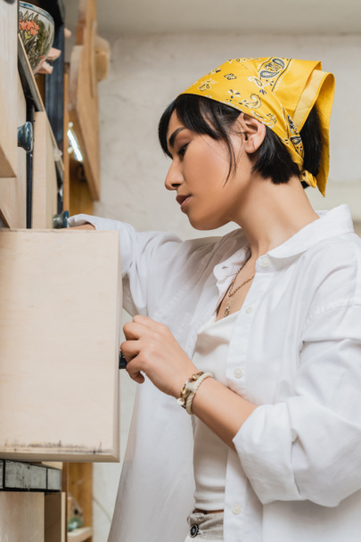 Seitenansicht der jungen brünetten asiatischen Künstlerin mit Kopftuch und Arbeitskleidung, die den Schrank öffnet, während sie im Töpferatelier arbeitet, Töpferwerkstatt mit qualifizierter Kunsthandwerkerin - Foto, Bild