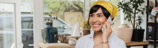 Lächelnde junge asiatische Kunsthandwerkerin in Arbeitskleidung und Kopftuch, die mit dem Smartphone spricht und in einer verschwommenen Keramikwerkstatt steht, Kunsthandwerkerin im Töpferatelier, die sich auf die Kreation konzentriert, Banner  - Foto, Bild