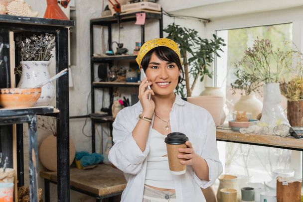 Türban ve iş kıyafetleri içinde neşeli genç Asyalı bayan çömlekçi akıllı telefonla konuşuyor ve kahve tutuyor bulanık çömlekçilik atölyesinde duruyor, çömlekçilik atölyesinde sanat eseri yaratmaya odaklanıyor. - Fotoğraf, Görsel