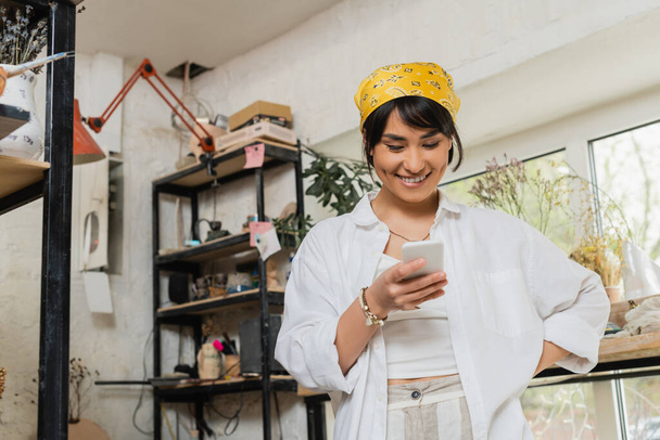 Θετική νεαρή ασιατική γυναίκα αγγειοπλάστη σε μαντίλα και ενδύματα εργασίας χρησιμοποιώντας smartphone, ενώ στέκεται και εργάζεται σε θολή κεραμικό εργαστήριο, τεχνίτης σε στούντιο κεραμικής με έμφαση στη δημιουργία - Φωτογραφία, εικόνα