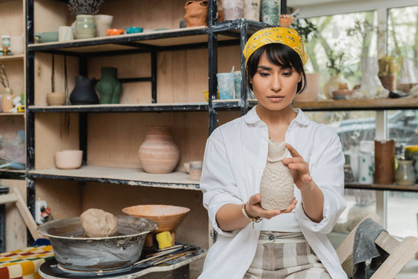 Jonge Aziatische brunette vrouwelijke pottenbakker in hoofddoek en werkkleding met klei sculptuur in de buurt van aardewerk wiel tijdens het werken in keramische werkplaats, vakmanschap in aardewerk maken - Foto, afbeelding