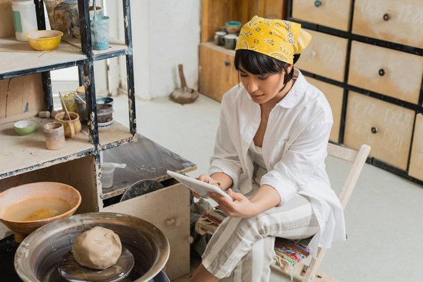 Jonge brunette Aziatische ambachtsman in hoofddoek en werkkleding met behulp van digitale tablet, terwijl zitten in de buurt van aardewerk wiel in wazig keramiek workshop, vakmanschap in aardewerk maken - Foto, afbeelding