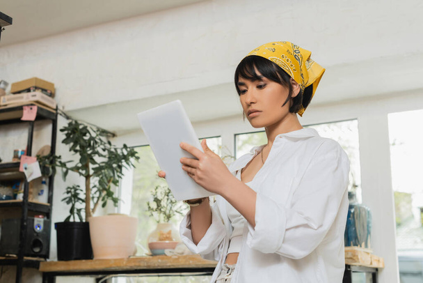 Young brunette asian female artist in headscarf and workwear using digital tablet while working in blurred ceramic workshop at background, craftsmanship in pottery making - Photo, Image