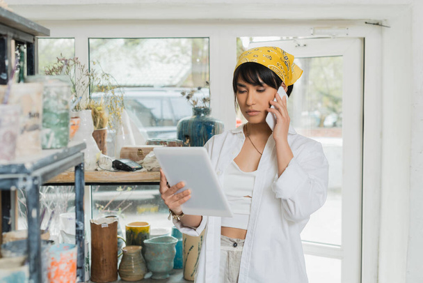 Nuori aasialainen naisartisti huivissa puhuu älypuhelimella ja käyttää digitaalista tablettia työskennellessään hämärtyneessä keraamisessa työpajassa taustalla, luova keramiikan valmistusprosessi - Valokuva, kuva