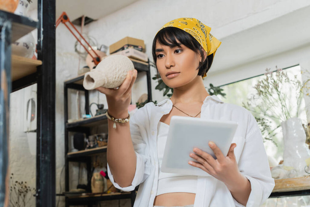 Junge asiatische brünette Kunsthandwerkerin in Arbeitskleidung und Kopftuch hält ein digitales Tablet in der Hand und betrachtet die Tonskulptur, während sie im Hintergrund in einer verschwommenen Keramikwerkstatt steht, Prozess der Töpferei - Foto, Bild