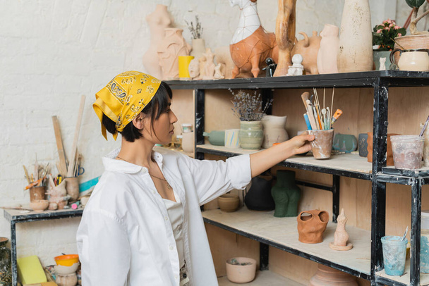 Widok z boku młodego azjatyckiego rzemieślnika w chustce i odzieży roboczej biorąc narzędzia ceramiczne z półki z wyrobów ceramicznych w studiu ceramicznym, proces twórczy ceramiki - Zdjęcie, obraz