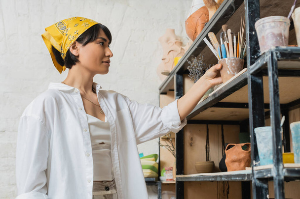 Junge asiatische Künstlerin in Arbeitskleidung und Kopftuch, die Töpferwerkzeuge ins Regal in der Nähe von Tonprodukten stellt und in einer Keramikwerkstatt arbeitet, kreativer Prozess der Töpferei - Foto, Bild