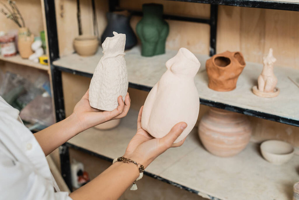 Cropped view of young female artisan in workwear holding clay sculptures while standing near blurred rack in ceramic workshop, pottery studio scene with skilled artisan - Fotoğraf, Görsel