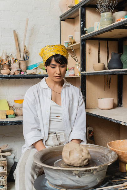 Young asian female artisan in headscarf and workwear looking at clay on pottery wheel while sitting near rack with sculptures in workshop, pottery studio scene with skilled artisan - Photo, Image