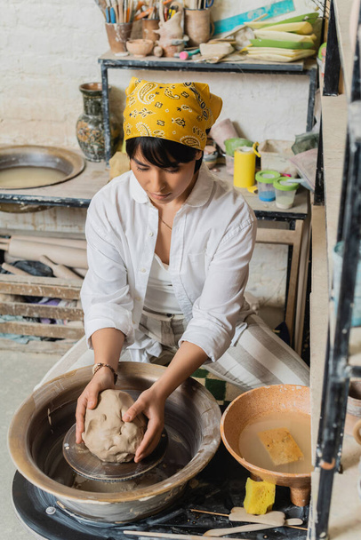Young brunette female potter in headscarf and workwear holding molding clay on pottery wheel while sitting near bowl with water in ceramic workshop, pottery studio scene with skilled artisan - Photo, Image