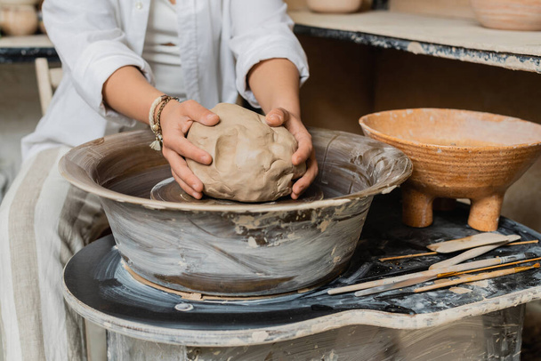 Vista ritagliata di donna sfocata artigiana in abbigliamento da lavoro mettendo argilla sulla ruota in ceramica vicino agli strumenti e ciotola in laboratorio di ceramica, scena in studio di ceramica con abile artigiano - Foto, immagini