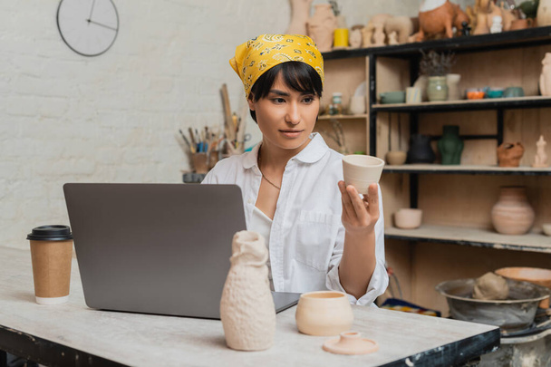 Молодая азиатская брюнетка ремесленница в рабочей одежде держит глиняный продукт возле ноутбука и кофе, чтобы пойти на стол в размытой керамической мастерской на заднем плане, керамика художник демонстрирует ремесло - Фото, изображение