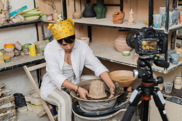 Jeune artisan asiatique en vêtements de travail et foulard moulant l'argile sur la roue de la poterie près de l'appareil photo numérique flou sur trépied dans l'atelier de céramique, artiste de la poterie mettant en valeur l'artisanat - Photo, image