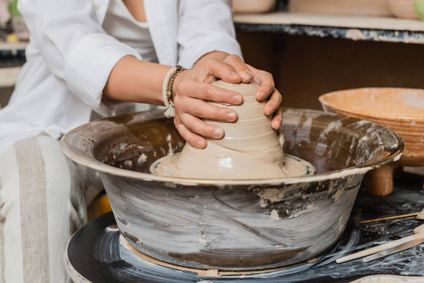 Ausgeschnittene Ansicht einer verschwommenen Handwerkerin in Arbeitskleidung, die nassen Ton auf Töpferscheibe in der Nähe von Schüssel im Hintergrund in Keramikwerkstatt, Töpferatelier und Bastelkonzept formt - Foto, Bild