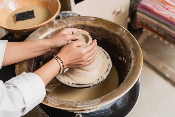 Cropped view of young craftswoman shaping wet clay on pottery wheel near blurred bowl with water and sponge in ceramic workshop, pottery studio workspace and craft concept - Photo, Image