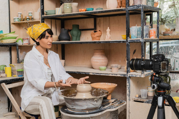 Ázsiai női kézműves fejkendő és munkaruha beszél a digitális fényképezőgép állvány, miközben dolgozik nedves agyag kerámia kerék közelében állvány művészeti műhely, agyag szobrászat folyamat koncepció - Fotó, kép