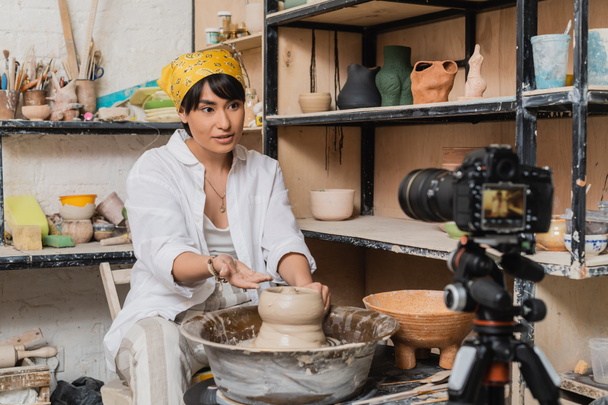 Asian female artisan in workwear and headscarf talking and gesturing at digital camera on tripod while working with clay on pottery wheel in art workshop, clay sculpting process concept - Photo, Image