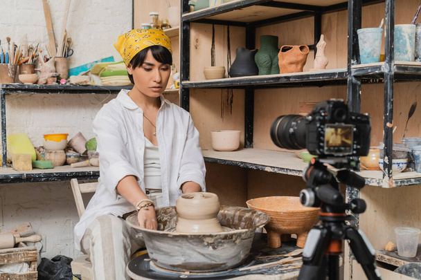Młody azjatycki artysta w chustce i odzieży roboczej, pracujący z mokrą gliną na kole garncarskim w pobliżu rozmytego aparatu cyfrowego na statywie w warsztacie ceramicznym, koncepcja procesu rzeźbienia gliny - Zdjęcie, obraz