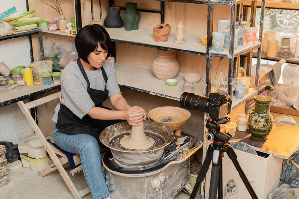 Young asian female artisan in apron shaping wet clay on pottery wheel near bowl with water and tools near digital camera in ceramic workshop at background, pottery tools and equipment - Photo, Image