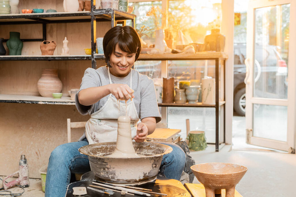 Улыбающаяся молодая азиатская художница в фартуке льет воду на глину на гончарное колесо рядом с инструментами и чашу в размытой мастерской на закате, ремесленник создает уникальные керамические изделия - Фото, изображение
