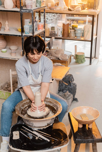 Young brunette asian artisan in apron molding wet clay on pottery wheel near bowl with water and tools in blurred ceramic workshop at sunset, artisan creating unique pottery pieces - Photo, Image