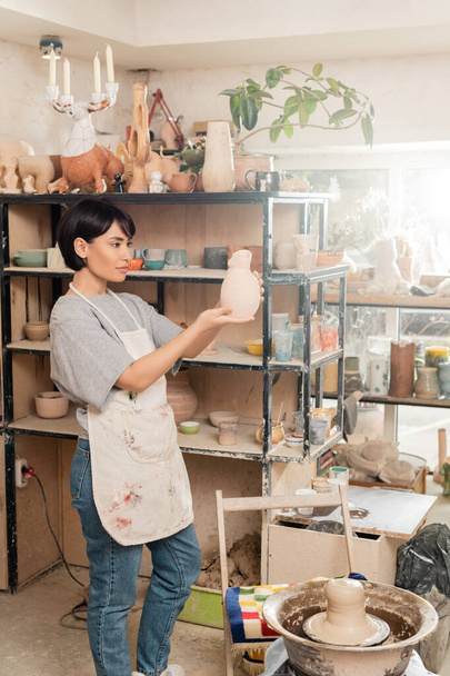 Young brunette asian female artist in apron holding sculpture while standing near clay on pottery wheel and rack in blurred ceramic studio at sunset, clay sculpting process concept - Photo, Image