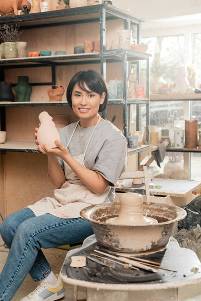 Sonriente joven asiática artesana en delantal sosteniendo escultura de cerámica y mirando a la cámara mientras está sentado cerca de arcilla húmeda en la rueda de cerámica en taller borroso, concepto de proceso de escultura de arcilla - Foto, Imagen