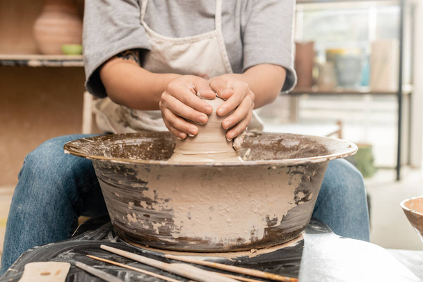 Oříznutý pohled na tetované ženské řemeslnice v zástěře tvarování mokré hlíny a práce s keramickým kolem v blízkosti rozmazané nástroje na stole v rozmazané keramické dílně, hliněné sochařství koncepce procesu - Fotografie, Obrázek
