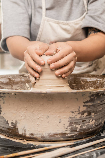 Widok plonu zamazane i wytatuowane rzemieślniczka w fartuchu formowania mokrej gliny podczas pracy na kole ceramiki w pobliżu narzędzi na stole w studiu ceramicznym, koncepcji procesu rzeźbienia gliny - Zdjęcie, obraz