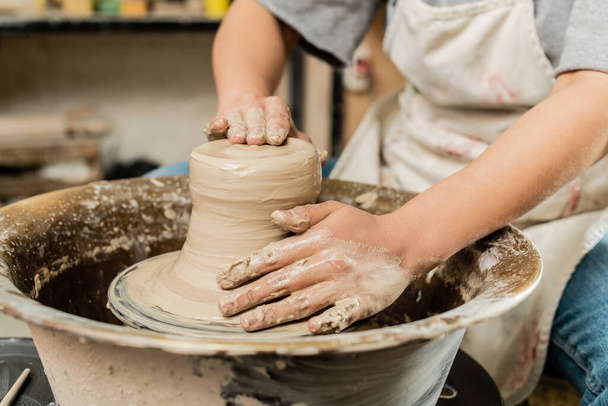 Vista ritagliata di sfocato femminile artigiano in grembiule modellare argilla bagnata e lavorare con ruota ceramica in laboratorio d'arte ceramica sullo sfondo, concetto di ceramica qualificato - Foto, immagini