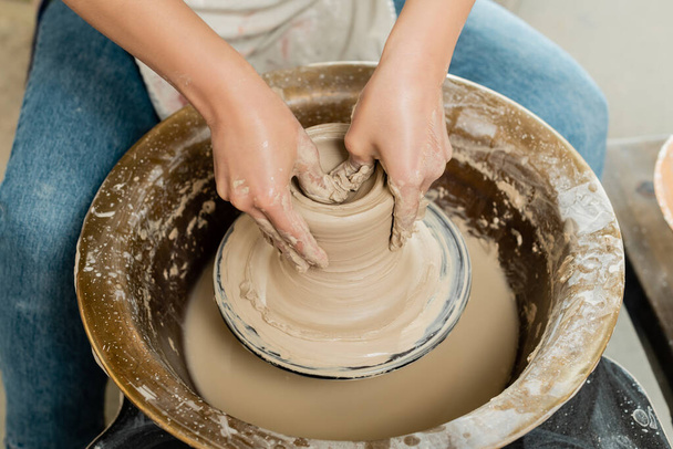 High angle view of young female potter in apron shaping wet clay while working on spinning pottery wheel in art workshop at background, skilled pottery making concept - Photo, Image