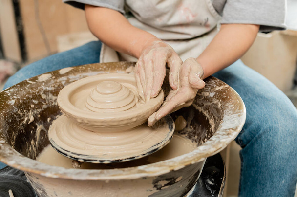 Cropped view of blurred female artisan in apron making shape of clay on spinning pottery wheel while working in pottery class, skilled pottery making concept - Photo, Image