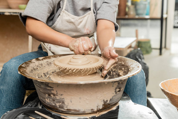Vista ritagliata del giovane artigiano femminile in grembiule versando acqua dalla spugna sull'argilla sulla ruota della ceramica di filatura vicino alla ciotola in studio d'arte sfocata, processo di creazione della ceramica - Foto, immagini